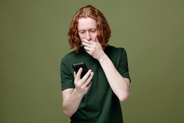 Bezpłatne zdjęcie przestraszone, zakryte usta ręką młody przystojny facet trzymający i patrzący na telefon ubrany w zieloną koszulkę na białym tle na zielonym tle