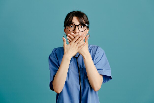 Przestraszone, zakryte usta rękami młoda lekarka nosi jednolity stetoskop fith na białym tle na niebieskim tle