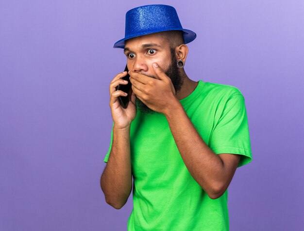 Przestraszony młody Afro-Amerykanin w kapeluszu imprezowym mówi przez telefon zakryty dłonią