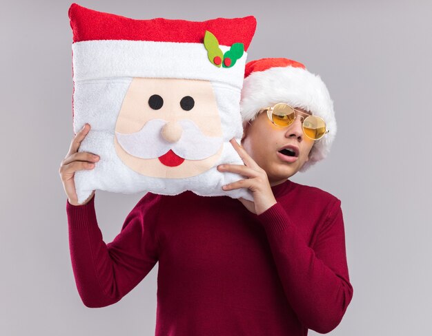 Przestraszony młody chłopak w kapeluszu Boże Narodzenie w okularach, trzymając poduszkę świąteczną na białym tle