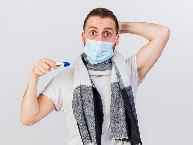Przestraszony młody chory człowiek ubrany w maskę medyczną i szalik, trzymając termometr i kładąc rękę za głowę na białym tle