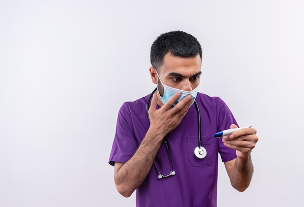 Przestraszony Młody Lekarz Mężczyzna Ubrany W Fioletową Odzież Chirurga I Maskę Medyczną Stetoskop Patrząc Na Termometr W Ręce Zakryte Usta Ręką Na Na Białym Tle