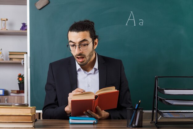 Przestraszony nauczyciel w okularach, trzymający i czytający książkę, siedzący przy stole z szkolnymi narzędziami w klasie