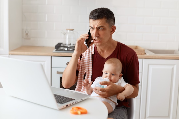 Przestraszony poważny mężczyzna ubrany w bordową casualową koszulkę z ręcznikiem na ramieniu, opiekujący się dzieckiem i pracujący online z domu, rozmawiający przez telefon z partnerem, mający kłopoty w pracy.