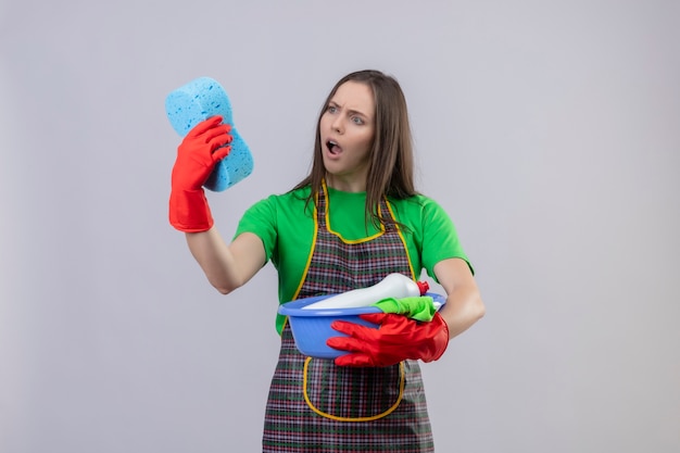 Przestraszony sprzątanie młoda dziewczyna ubrana w mundur w czerwonych rękawiczkach trzymając narzędzia do czyszczenia, patrząc na gąbkę na dłoni na na białym tle