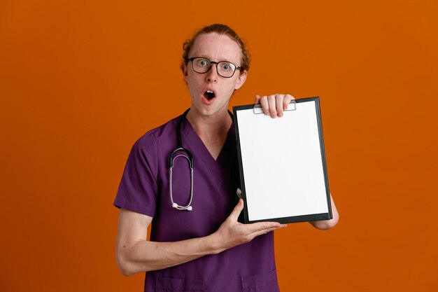 Przestraszony, trzymając schowek, młody mężczyzna lekarz ubrany w mundur ze stetoskopem na białym tle na pomarańczowym tle