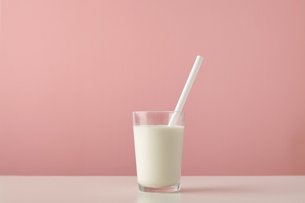 Przezroczyste szkło ze świeżym organicznym mlekiem i białą słomką wewnątrz na białym tle na pastelowym różowym tle na drewnianym stole