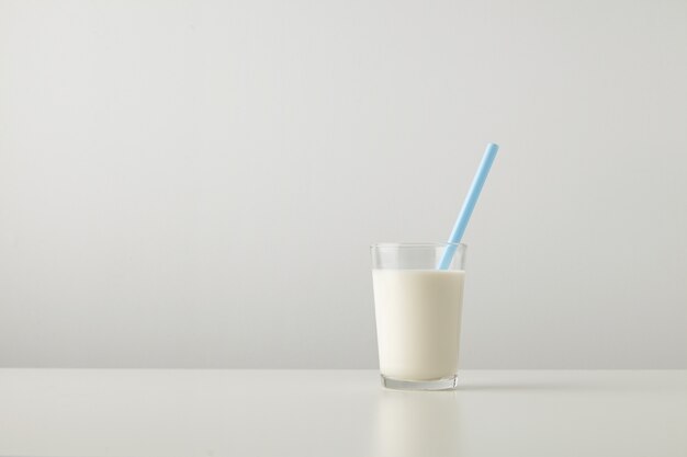 Przezroczyste szkło ze świeżym organicznym mlekiem i niebieską słomką wewnątrz na białym tle z boku białego stołu