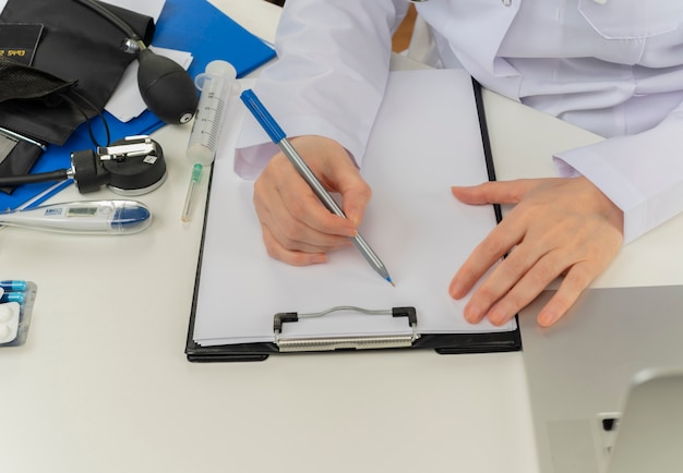 Przycięte strzał kobiece ręce lekarza pracującego przy biurku z narzędzi medycznych i laptopa, pisząc receptę w schowku
