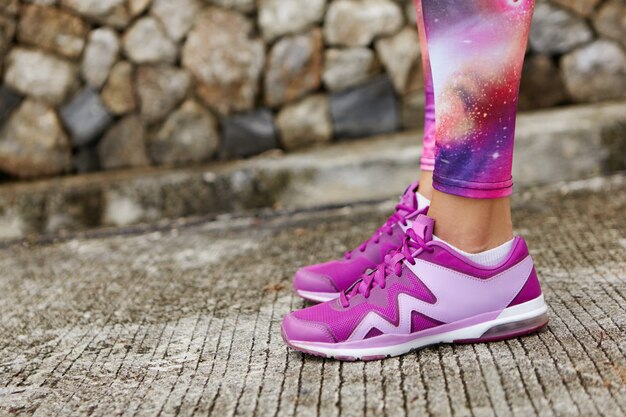 Przycięte ujęcie sprawnej biegaczki w fioletowych butach do biegania i legginsach z nadrukiem kosmicznym, stojącej na kamiennym betonie, przygotowując się do treningu do biegania.