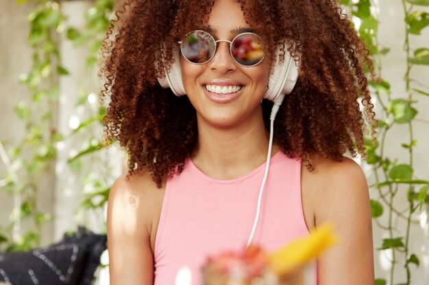 Przycięte ujęcie zadowolonej ciemnoskórej kobiety z krzaczastą fryzurą w stylu afro słucha ulubionej playlisty w słuchawkach, jest w dobrym nastroju, nosi modne okulary przeciwsłoneczne i różową koszulkę, ma promienny uśmiech