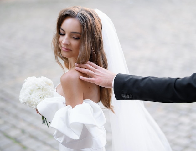 Bezpłatne zdjęcie przycięte uroczej panny młodej z długim welonem ubranej w suknię ślubną z bukietem białych kwiatów i patrzącą za siebie, podczas gdy ręka nierozpoznawalnego mężczyzny dotyka jej nagiego ramienia na ulicy