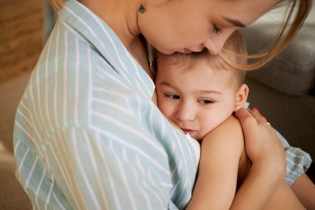 Przycięty obraz czułej, troskliwej młodej matki tulącej senne dziecko w ramionach, śpiewającej mu piosenkę i całującej się w czoło. Mama przytulająca roczne małe dziecko, próbująca złagodzić skurcze brzucha