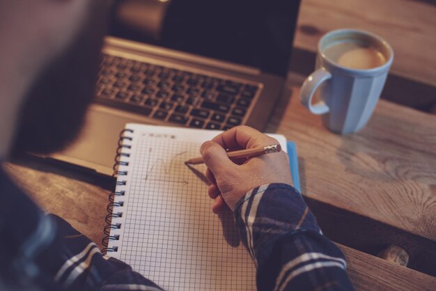 Przycięty obraz przypadkowego biznesmena lub freelancera planującego pracę na notebooku, pracującego na laptopie ze smartfonem, filiżankę kawy na stole w kawiarni lub biurze domowym, pracującego z kawiarni c