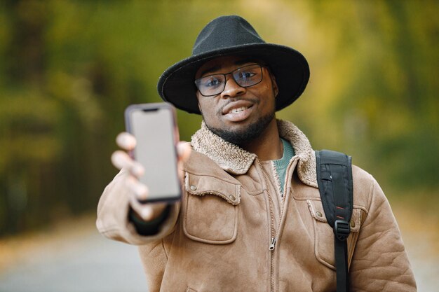 Przycięty portret czarnego podróżnika męskiego pokazujący pusty ekran telefonu komórkowego dla informacji lub reklamy. Selektywne skupienie. Mężczyzna stojący na zewnątrz i ubrany w brązową kurtkę, okulary i czerń