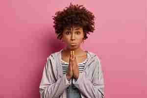 Bezpłatne zdjęcie przygnębiona nerwowa etniczna kobieta z kręconymi włosami, zdenerwowana błagalnym grymasem, modli się, błaga, zaciska usta, nosi zwykłą kurtkę, pozuje na różowej ścianie, błaga z nadzieją