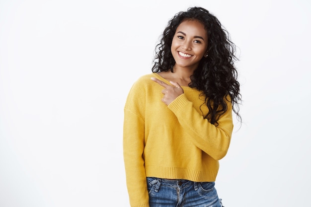 Przyjazna atrakcyjna afroamerykańska kobieta w żółtym stylowym swetrze uśmiecha się zadowolona, wygląda na pewną siebie i beztroską, dzieli się fajnymi wiadomościami, wskazuje w lewo, daje radę sprawdź fajny sklep