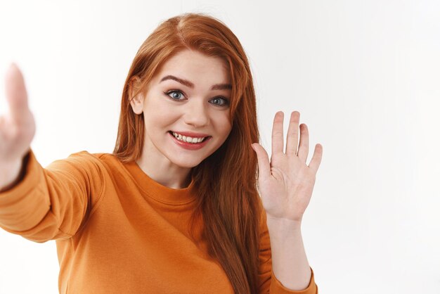 Przyjazna i otwarta ładna rudowłosa kobieta w pomarańczowym swetrze wysuwa rękę trzymającą smartfona lub kamerę, biorąc selfie machając ręką w cześć lub cześć uśmiechnięta znajoma rozmawiająca wideo za pomocą aplikacji online