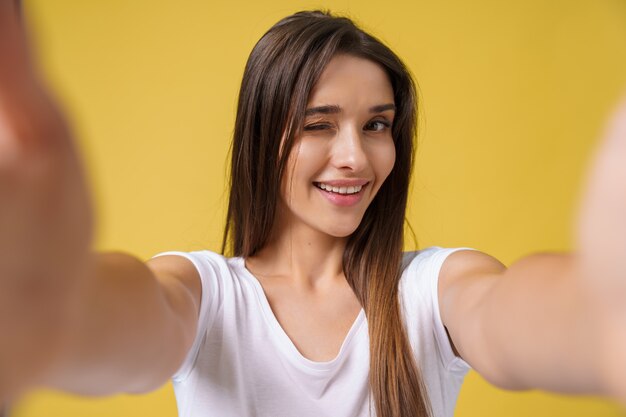 Przyjemna atrakcyjna dziewczyna co selfie w studio i śmiejąc się. Przystojna młoda kobieta z brązowymi włosami robi sobie zdjęcie na jasnożółtym tle.
