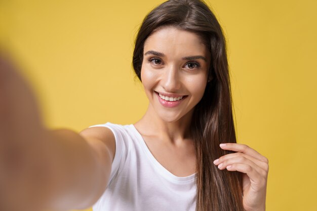 Przyjemna atrakcyjna dziewczyna co selfie w studio i śmiejąc się. Przystojna młoda kobieta z brązowymi włosami robi sobie zdjęcie na jasnożółtym tle.