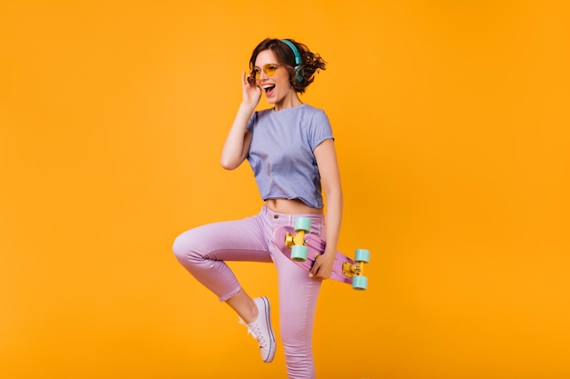 Przyjemna kaukaska dziewczyna w różowych obcisłych spodniach tańcząca na pomarańczowym tle Studio fotografii kręconej damy w słuchawkach pozującej z longboardem i śmiejącej się
