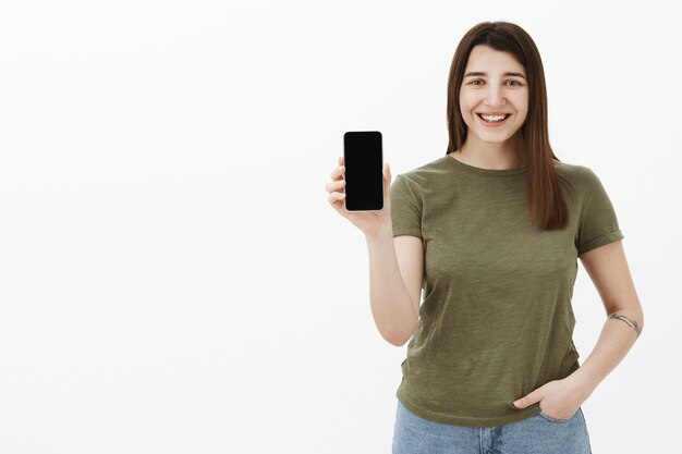 Przyjemnie wyglądająca, radosna i beztroska młoda brunetka z lat dwudziestych z tatuażem w luźnej oliwkowej koszulce, uśmiechnięta i śmiejąca się czule, jak trzyma smartfon prezentujący aplikację na ekranie telefonu komórkowego nad szarą ścianą