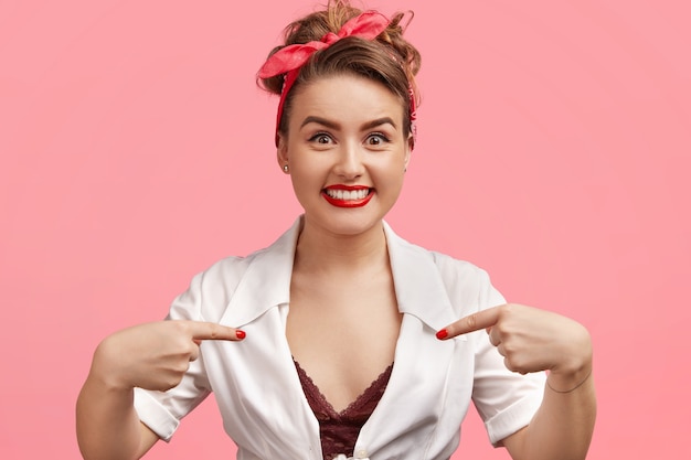Bezpłatne zdjęcie przyjemnie wyglądająca, szczęśliwa młoda kobieta nosi stylową czerwoną opaskę i białą koszulę, wskazuje na siebie dwoma palcami wskazującymi