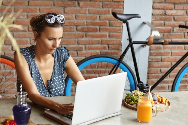 Przypadkowa bizneswoman nosi okulary przeciwsłoneczne na głowie i gra na klawiaturze laptopa, sprawdzając e-maile podczas lunchu w jej dzień wolny