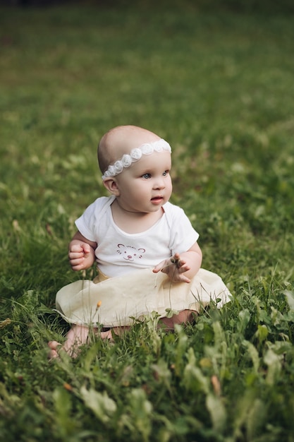 Przystojna mała dziewczynka z krótkimi jasnymi włosami i ładnym uśmiechem w białej sukni siedzi na trawie w parku latem i uśmiecha się
