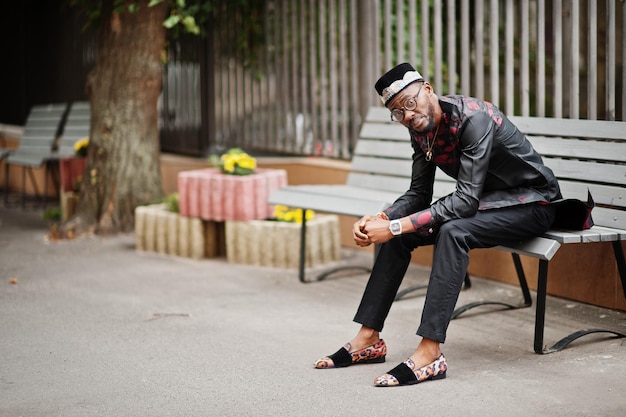 Przystojny afro amerykański mężczyzna w tradycyjnej czapce i okularach w nowoczesnym mieście siedzący na ławce
