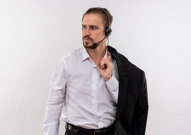 Przystojny biznesmen trzymając kurtkę na ramieniu ze słuchawkami z mikrofonem patrząc na bok z poważną twarzą stojącą na białym tle