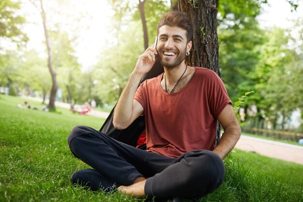 Przystojny brodaty facet odpoczywa w parku na trawie, rozmawia przez telefon komórkowy i uśmiecha się szczęśliwy