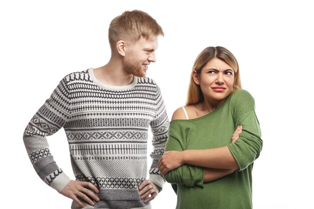 Przystojny brodaty facet ubrany w sweter uśmiecha się i patrzy na atrakcyjną kobietę, która stoi w zamkniętej pozycji z założonymi rękami, czuje się zdezorientowana, ponieważ nie lubi lub nie rozumie jego głupiego żartu