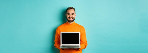 Przystojny brodaty mężczyzna w pomarańczowym swetrze pokazujący ekran laptopa demonstrujący stojący sklep internetowy