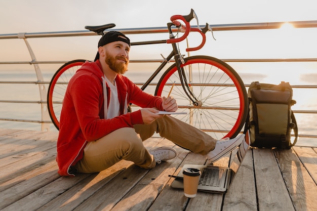 Przystojny Brodaty Mężczyzna W Stylu Hipster Pracujący Online Freelancer Na Laptopie Z Plecakiem I Rowerem W Porannym Wschodzie Słońca Nad Morzem Zdrowy Aktywny Styl życia Podróżnik Z Plecakiem