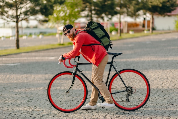Przystojny brodaty mężczyzna w stylu hipster w czerwonej bluzie z kapturem i okularach przeciwsłonecznych, jeżdżący samotnie z plecakiem na rowerze, podróżujący z plecakiem zdrowego, aktywnego stylu życia