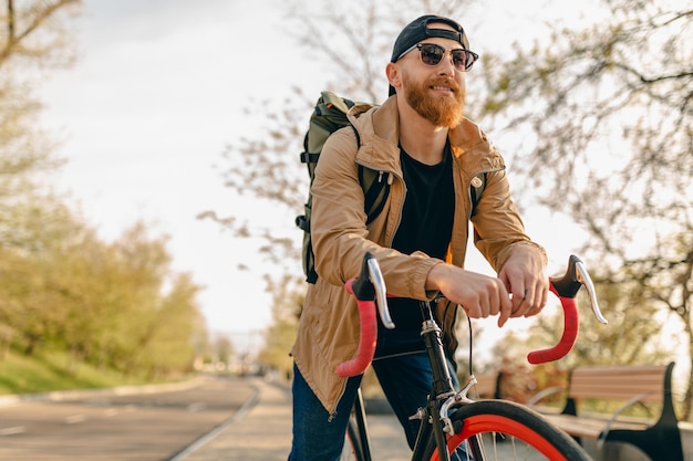 Przystojny brodaty mężczyzna w stylu hipster w kurtce i okularach przeciwsłonecznych, jeżdżący sam z plecakiem na rowerze, podróżnik zdrowego, aktywnego stylu życia