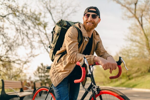 Przystojny brodaty mężczyzna w stylu hipster w kurtce i okularach przeciwsłonecznych, jeżdżący sam z plecakiem na rowerze, podróżnik zdrowego, aktywnego stylu życia