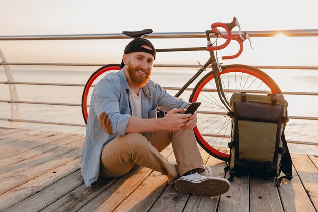 Przystojny brodaty mężczyzna w stylu hipster z plecakiem trzymając smartfona w dżinsowej koszuli i czapce z rowerem w porannym wschodzie słońca nad morzem zdrowy aktywny styl życia podróżnik z plecakiem