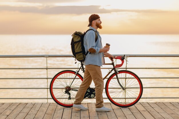 Przystojny brodaty mężczyzna w stylu hipster z plecakiem w dżinsowej koszuli i czapce z rowerem o porannym wschodzie słońca nad morzem pije kawę, zdrowy, aktywny styl życia podróżnik z plecakiem