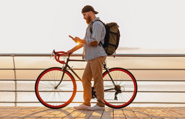 Przystojny brodaty mężczyzna w stylu hipster z plecakiem w dżinsowej koszuli i czapce z rowerem o porannym wschodzie słońca nad morzem pije kawę, zdrowy, aktywny styl życia podróżnik z plecakiem