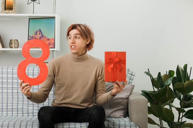 Przystojny facet na szczęśliwy dzień kobiet trzymający numer osiem z obecnym siedzącym na kanapie w salonie