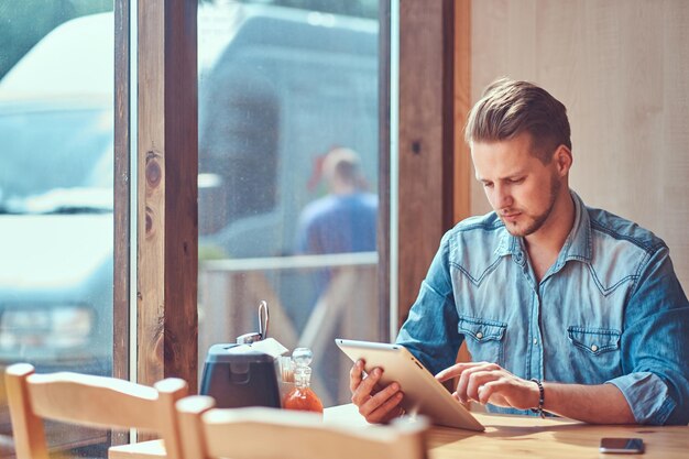 Przystojny hipster ze stylową fryzurą i brodą siedzi przy stoliku w przydrożnej kawiarni, wygląda coś na tablecie.