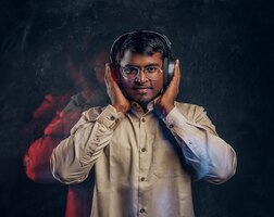 Bezpłatne zdjęcie przystojny indianin cieszący się słuchaniem muzyki w słuchawkach bezprzewodowych stojący w studio na tle ciemnej ściany