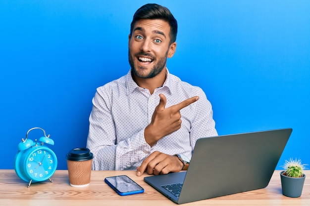 Przystojny latynos pracujący przy użyciu laptopa w biurze uśmiecha się wesoło, wskazując ręką i palcem na bok