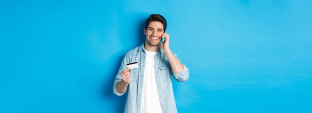 Przystojny Mężczyzna Dzwoni Do Banku I Trzyma Kartę Kredytową, Rozmawiając Przez Telefon Komórkowy Stojąc Nad Niebieskim Tyłem