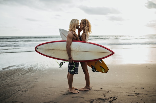 Przystojny mężczyzna i jego dziewczyna całują się na tle morza i trzymając deski surfingowe