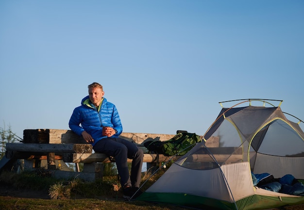 Bezpłatne zdjęcie przystojny mężczyzna podróżnik siedzący na ławce w pobliżu namiotu obozowego