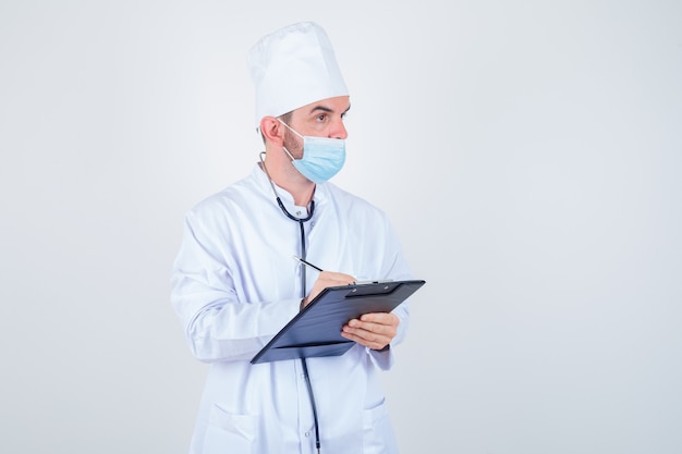 Przystojny mężczyzna trzyma stetoskop na szyi, pisze notatki w schowku w białym fartuchu medycznym, masce i zamyślony, widok z przodu.