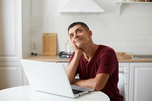 Przystojny mężczyzna ubrany w bordową koszulkę w stylu casual, siedzący przed komputerem przenośnym przy stole w kuchni, marzący o wakacjach, wyobraża sobie, jak spędzi weekend.